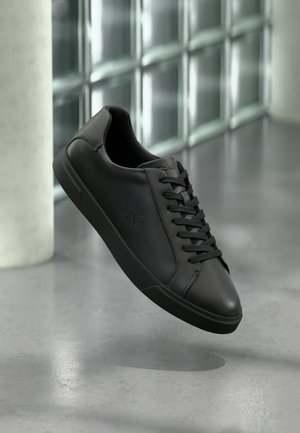 Baskets en cuir noir avec lacets et logo "ck" en relief flottant au-dessus d'un sol gris près d'un mur en verre brique.