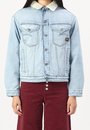 Vrouw draagt een lichtblauwe spijkerjassen met een witte fleece kraag en bordeauxrode, hooggetailleerde broek met drie zichtbare knopen.