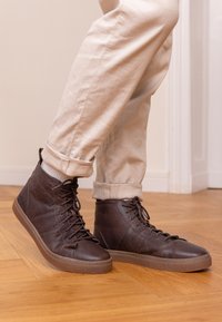 Braune Leder-Hochtop-Sneaker mit strukturierter Oberfläche, ausrüstet mit Schnürdesign und Gummisohle, kombiniert mit cremefarbenen Pants.