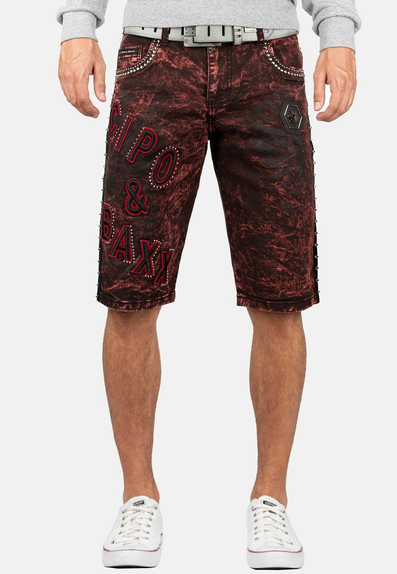Shorts en denim rouge et noir avec un effet vieilli, présentant un grand texte de marque brodé, des clous en métal et des boutons sur les côtés pour ajouter des détails.