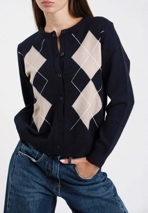 Donna che indossa un cardigan blu navy con motivo beige a rombi e bottoni neri, abbinato a jeans blu a vita alta, mano in tasca.