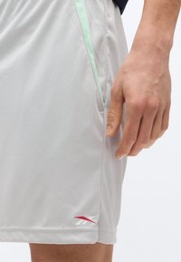 Κοντινή λήψη του χεριού ενός ατόμου κοντά στην πλαϊνή τσέπη ενός ανοιχτόχρωμου γκρι αθλητικού σορτς με κόκκινο και άσπρο λογότυπο Reebok.