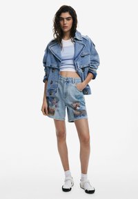 Denimtakki, jossa on sinertävä sävy, kaksirivinen rakenne ja voimakkaat kaulukset. Yhdistetty vaaleansinisiin shortseihin, joissa on värikkäitä printtejä.