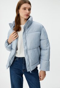 Mujer con chaqueta de plumas azul clara, camisa blanca y jeans azules, sosteniendo el cuello de la chaqueta, mirando de lado frente a un fondo liso.