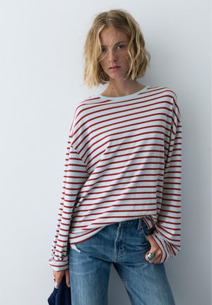 Blondínka nosiaca voľný biely sveter s červenými horizontálnymi pruhmi, modré džíny a drží v ľavej ruke tmavomodrú látku.