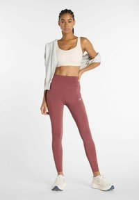 Leggings burdeos con cintura alta, combinadas con un sujetador deportivo beige y un suéter gris claro colgado sobre un hombro. Zapatillas blancas.