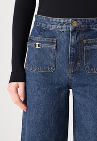 Mörkblå vida denimjeans med guldverktyg, två bakfickor och orange sytråd. Har hög midja och en strukturerad yta.