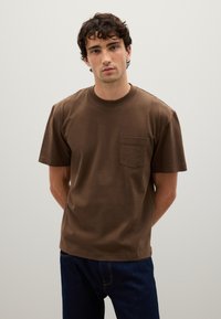 T-shirt in cotone marrone con scollatura rotonda e maniche corte. Presenta una tasca sul petto e una vestibilità comoda. Indossato con jeans blu.