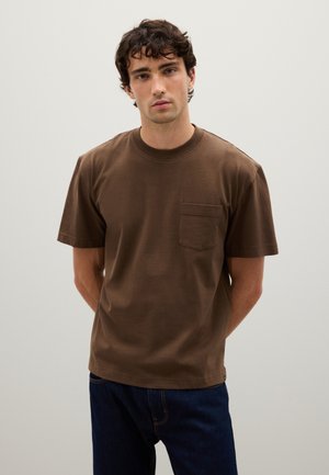 Camiseta de algodón marrón con cuello redondo y mangas cortas. Cuenta con un bolsillo en el pecho y un corte holgado. Llevada con jeans azules.