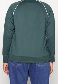 Mörkgrön sweatshirt med vit kant, ribbade manschetter och nederkant, slät textur och avslappnad passform, tillsammans med blå denimbyxor.