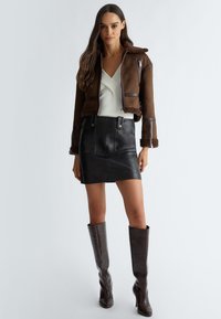 Giacca cropped in suede marrone con rifiniture in shearling, abbinata a una mini gonna in pelle nera. Stivali marroni al ginocchio in textured completano il look.
