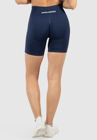 Model in marineblauwe sportshorts en witte sneakers, gezien van achteren, die de pasvorm van de shorts en het merkembleem op de tailleband toont.