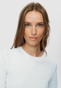 Mlada žena s dugom smešnom kosom i svijetloplavim očima, obučena u bijelu majicu, gleda izravno u kameru s neutralnim izrazom lica.