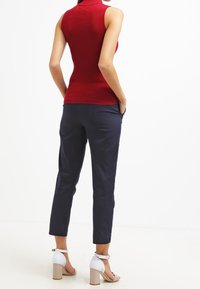 Vrouw met handen in haar zakken, gekleed in een mouwloze rode top, marineblauwe broek en witte blokhak sandalen op een witte achtergrond.