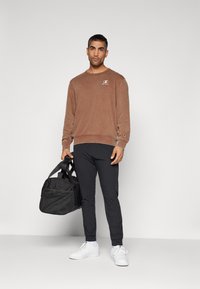 Felpa marrone in peluche con logo sul petto, abbinata a pantaloni neri slim-fit e sneakers bianche. Tiene una borsa a tracolla nera.
