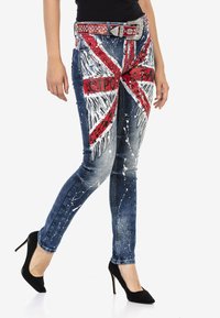 Verblasste blaue Jeans mit einem Union Jack-Muster in Rot und Weiß, versehen mit Spritzereien, kombiniert mit einem besetzten Gürtel und schwarzen Absatzschuhen.