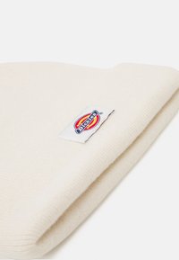 Cream-colored knitted beanie with a folded brim displaying a rectangular label featuring a red and yellow logo on a white background.