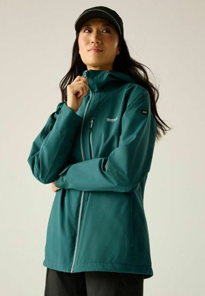 Vrouw draagt een teal Regatta outdoor jas, trekt de rits omhoog, met lang donker haar en een zwarte pet, tegen een effen achtergrond.