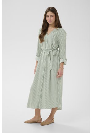 Mladá žena s dlhými hnedými vlasmi, oblečená v zeleno-bielych pruhovaných šatách na gombíky so zaväzovaním v páse a hnedými pletenými plochými topánkami.