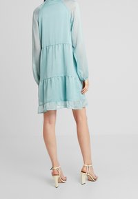 Une robe à volants bleu clair à pois, avec des manches semi-transparentes et un ourlet volanté, associée à des sandales à talons hauts beiges.