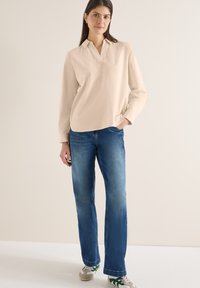 Femme debout, une main dans la poche, portant une chemise beige à manches longues, un jean bleu et des baskets blanches avec des rayures vertes.