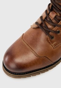 Barna bőr bokacsizma lekerekített orral, textúrált felülettel, barna fűzővel, fém szemekkel és markáns mintázatú gumitalppal.