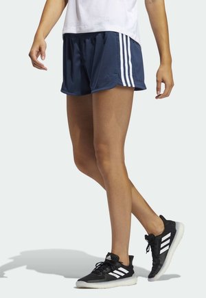 Persoon die een donkerblauwe Adidas short met witte strepen draagt en zwarte Adidas sneakers, staand met één voet licht opgetild.