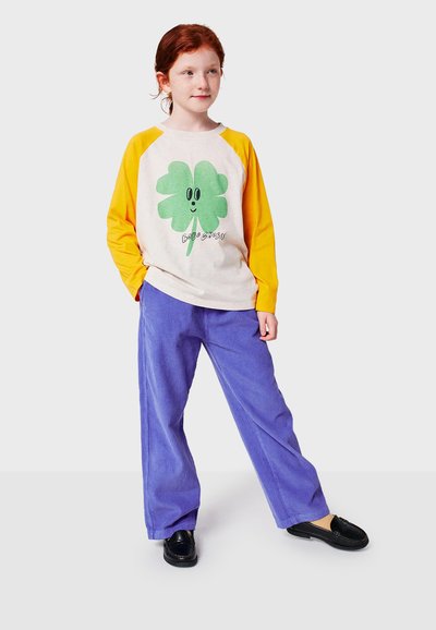 Long-sleeve, cream and yellow raglan shirt with green clover graphic, paired with purple corduroy wide-leg pants and black slip-on shoes.