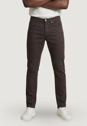 Hombre con pantalones ajustados de color marrón oscuro y zapatillas blancas, de pie con los brazos cruzados frente a un fondo claro y liso.