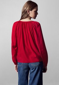 Blusa rossa a maniche lunghe con collo arricciato, texture liscia e vestibilità rilassata, abbinata a jeans in denim blu scuro.