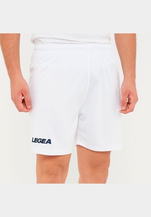 Mężczyzna ubrany w białe sportowe spodenki z elastycznym pasem i logo "LEGEA" na lewej nodze, stoi z ramionami swobodnie opuszczonymi wzdłuż ciała.