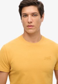 Camiseta de algodón amarilla con cuello redondo, mangas cortas y detalle de logo bordado en el lado izquierdo del pecho. Textura suave y diseño ajustado.