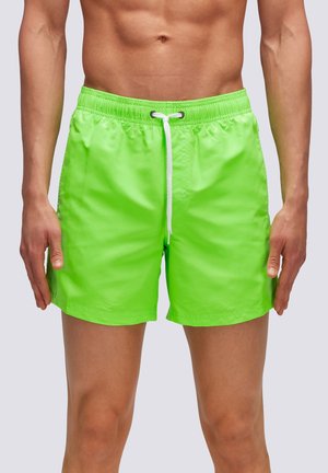 Torace maschile con pantaloncini da bagno verde brillante con coulisse bianca, braccia rilassate ai lati, sfondo chiaro e uniforme.