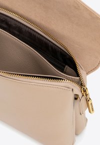 Beige kožená taška s texturovaným semišovým klopou a zlatým zipem, která obsahuje malý přívěsek. Dvě přihrádky s tmavým podšíváním.