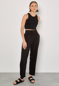 Schwarzes, kurzes Tanktop kombiniert mit lockeren schwarzen Hosen mit Kordelzug. Das Modell trägt schwarze Riemchensandalen mit Schnallen.