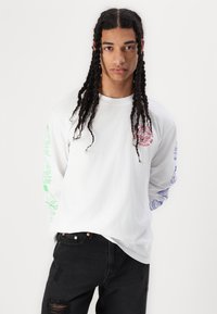Langærmet hvid t-shirt med grafiske print i grøn, rød og blå på ærmerne. Sort slidte jeans båret nedenfor. Standard pasform.