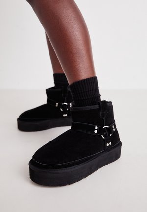 Persona con botines de ante negro con suelas gruesas y detalles de hebilla plateada, combinados con calcetines negros acanalados sobre un piso claro.