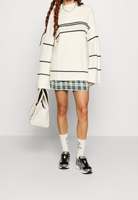 Suéter crema de gran tamaño con rayas negras, combinado con una falda corta de cuadros verdes. Los accesorios incluyen un bolso beige y calcetines blancos con diseños.