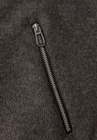 Reißverschlussdetails auf einem dunkelgrauen Filzstoff. Metallzähne und Schiebemechanismus sichtbar. Weiche Textur mit leichtem Glanz, die das Handwerk zeigt.