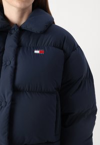 Marine pufferjack met een grote kraag, voorzien van een voorzak en een klein logo in rood, wit en blauw op de borst. Glad textuur.