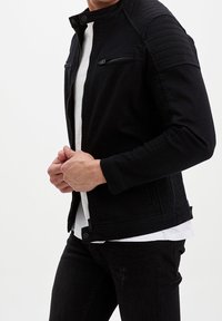 Czarna kurtka dżinsowa z usztywnianym kołnierzem, kieszeniami na zamek oraz pikowanymi detalami na ramionach. Noszona na białej koszulce.