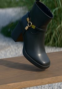 Black leather ankle boot with a chunky heel, featuring a gold ring and square accents on the strap. Smooth texture and sleek design.