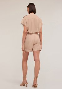 Blusa beige a maniche corte con vestibilità rilassata e piega sul retro. Abbinata a pantaloni corti beige a vita alta. Tacchi con cinturini avvolgenti.