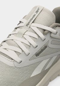 Μπεζ αθλητικό παπούτσι Reebok με άνω μέρος από δίχτυ, λευκά κορδόνια, υφασμάτινη λωρίδα με λογότυπο στη γλώσσα και λογότυπο Reebok στην πλευρική επιφάνεια.