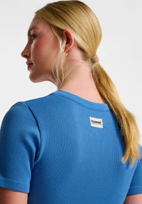 Niebieska prążkowana koszulka z krótkim rękawem, z okrągłym dekoltem oraz tkanym metkiem na plecach z napisem "hummel". Gładka tekstura.