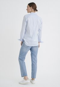 Light blue button-up shirt with long sleeves, paired with light blue denim jeans and white sneakers, seen from the back against a plain background.