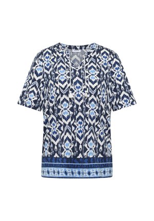 Blouse à manches courtes avec motifs géométriques et floraux bleus, blancs et noirs, col en V avec liens à pompons et bordure décorative à l'ourlet.