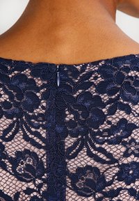 Marinblå spetsklänning som visar blommönster, intrikata detaljer och en dold dragkedja i ryggen. Halvgenomskinlig överdel med fast foder under.