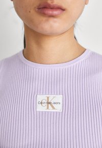 Ljubičasti rebrasti top s okruglim izrezom, koji sadrži bijelu etiketu s zlatnim natpisom "Calvin Klein Jeans" i stiliziranim "CK."