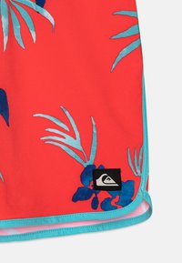 Röd tyg med blå och teal tropiska bladmönster, turkos kant och en svart Quiksilver-logopatch nära nederkanten.
